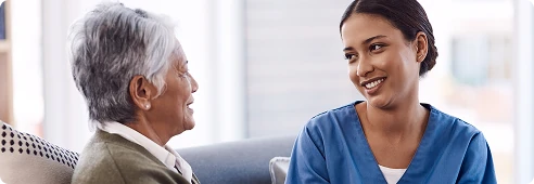 Caregiver in scrubs sitting with an older woman, smiling and engaging in friendly conversation.
