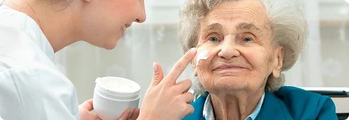 Caregiver applying cream to an elderly woman’s face, assisting with personal care needs.