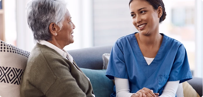 Caregiver in scrubs sitting on a couch, warmly talking with an elderly woman in a home setting.