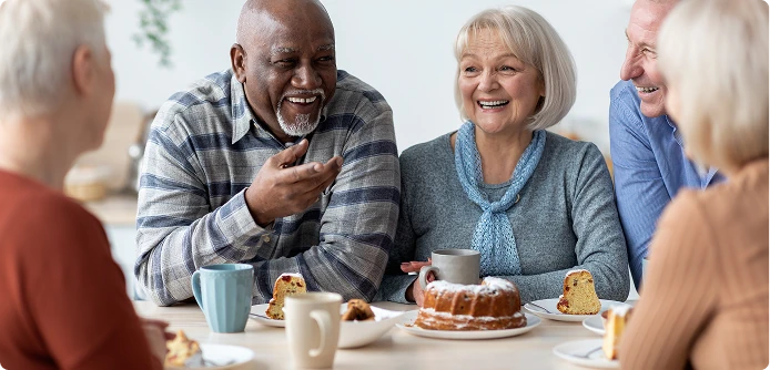 Group of older adults sitting around a table sharing coffee and cake, smiling and chatting together.